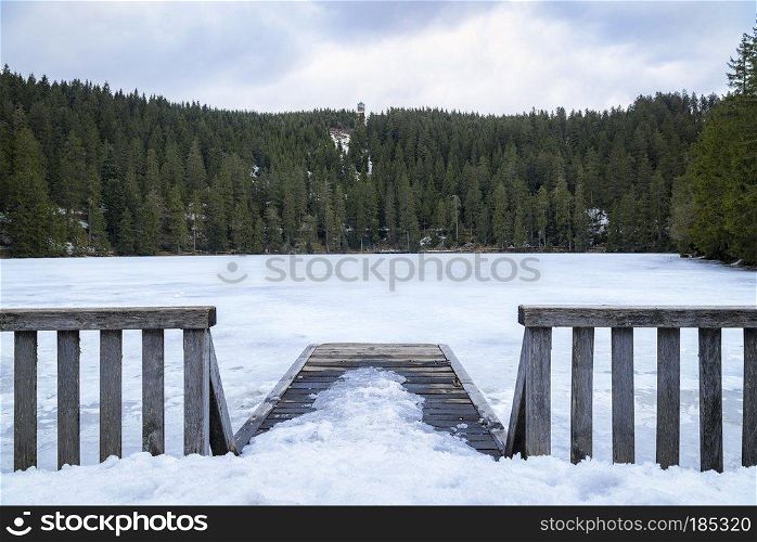 Landscape with snowy wooden bridge and the frozen water of the Mummelsee lake, surrounded by fir forest, in the Black Forest mountains, in Germany.