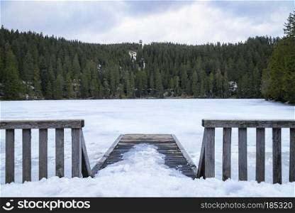 Landscape with snowy wooden bridge and the frozen water of the Mummelsee lake, surrounded by fir forest, in the Black Forest mountains, in Germany.