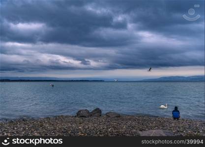 Landscape with the Bodensee lake, from in Friedrichshafen town, Germany, on a rainy day, while a man sits on its shore, meditating.