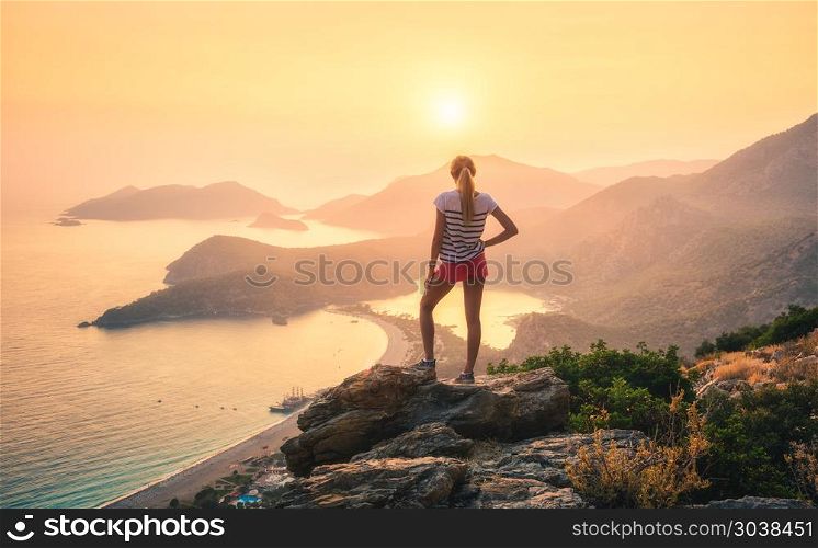 Landscape with woman, sea, mountain ridges and orange sky. Young woman standing on the top of rock and looking at the seashore and mountains at colorful sunset in summer. Landscape with girl, sea, mountain ridges and orange sunlight. Travel. Oludeniz, Turkey