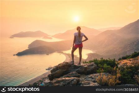 Landscape with woman, sea, mountain ridges and orange sky. Young woman standing on the top of rock and looking at the seashore and mountains at colorful sunset in summer. Landscape with girl, sea, mountain ridges and orange sunlight. Travel. Oludeniz, Turkey