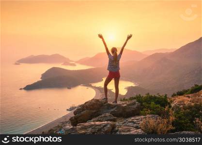 Landscape with woman, sea, mountain ridges and orange sky. Young woman with raised up arms standing on the top of rock and looking at the seashore and mountains at sunset. Summer landscape with girl, sea, islands and orange sunlight. Travel. Oludeniz, Turkey