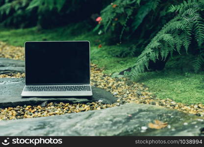 Laptop computer mockup has empty screen outside on stone in garden of coffee shop.