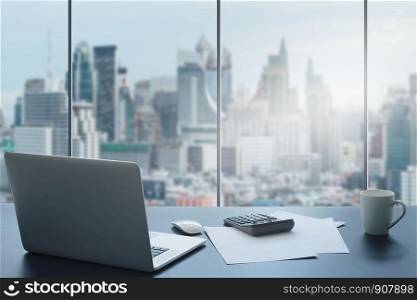 Laptop on table in office room on window city background.Computer and white paper,calculator with a coffee cup on a black desk