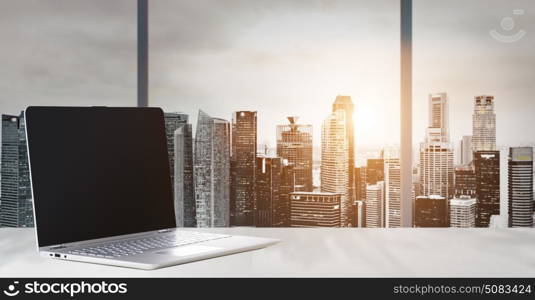 Laptop on table in office with panoramic sunset view of modern downtown skyscrapers at business district, blank screen