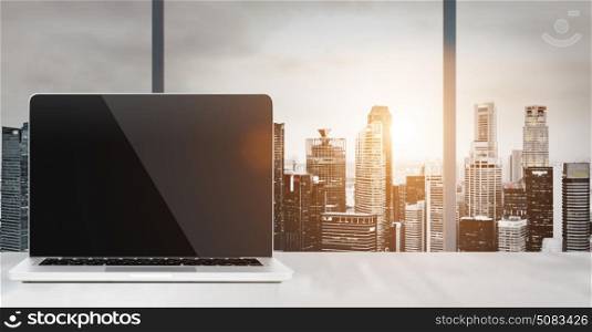 Laptop on table in office with panoramic sunset view of modern downtown skyscrapers at business district, blank screen