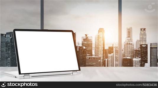 Laptop on table in office with panoramic sunset view of modern downtown skyscrapers at business district, blank screen