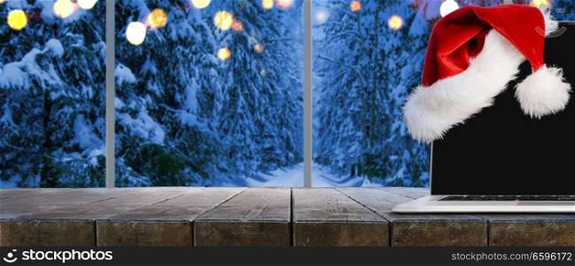 Laptop on table with Santa Claus hat at home with panoramic view through window of snowy trees in winter forest. Christmas work place with panoramic view