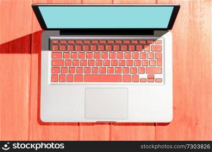 Laptop with living coral colored keyboard on wooden table on a sunny day, view from above. Laptop on wooden table