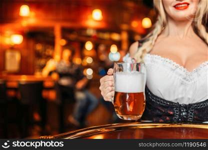 Large breasted waitress holds mug of fresh beer in pub. Octoberfest barmaid with attractive shapes in traditional style dress. Seductive woman serving table in bar