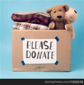 large brown cardboard box with the inscription please donate, box is filled with various clothes, toys on a blue background