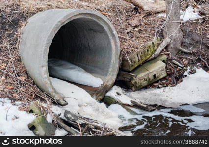 Large cement sewer drain pipe emptying into foamy puddle with debris in winter, surrounded by branches, dirt, and patches of snow.