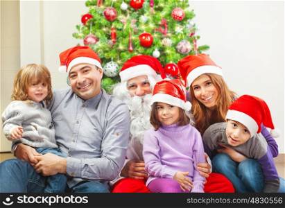 Large family and Santa Claus sitting near Christmas tree at home, wearing red festive hat, winter holidays, New Year celebration