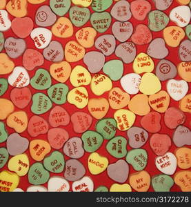 Large group of colorful candy hearts with sayings on them arranged on red background.