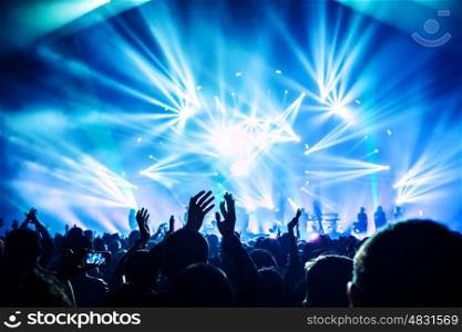 Large group of happy people enjoying rock concert, clapping with raised up hands, blue lights from the stage, new year celebration concept