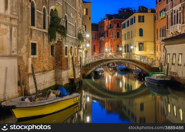 Lateral canal and pedestrian bridge in Venice at night with street light illuminating bridge and houses, with docked boats, Italy