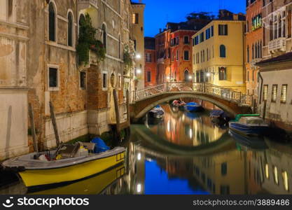 Lateral canal and pedestrian bridge in Venice at night with street light illuminating bridge and houses, with docked boats, Italy