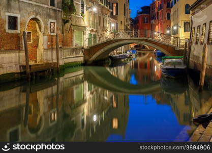 Lateral canal and pedestrian bridge in Venice at night with street light illuminating bridge and houses, with docked boats, Italy