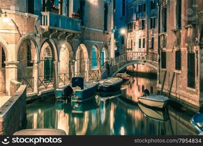 Lateral canal and pedestrian bridge in Venice at night with street light illuminating bridge and houses, with docked boats, Italy. Toning in cool tones