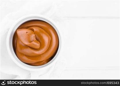 Latin American sweet caramel-like Manjar or Dulce de leche used as spread or filling in baking, photographed on white wood with copy space (Selective Focus, Focus on the top of the cream) . Latin American Manjar or Dulce de Leche