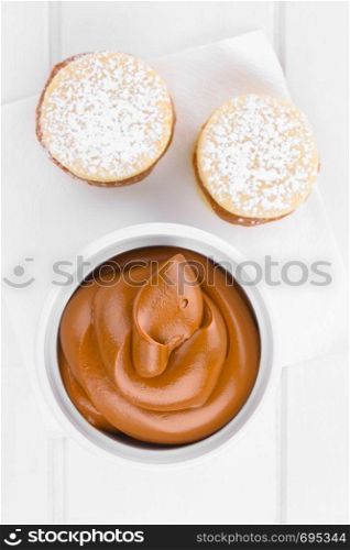 Latin American sweet caramel-like Manjar or Dulce de leche used as spread or filling in baking with alfajor cookies on the side, photographed overhead on white wood (Selective Focus, Focus on the top of the cream) . Latin American Manjar or Dulce de Leche