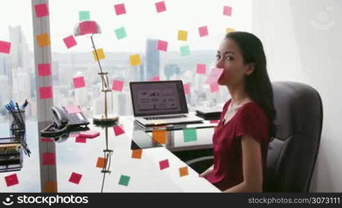 Latin american woman working as secretary in modern office with beautiful sight on the city, sticking adhesive notes with tasks on skyscraper window. The girl feels stressed, holding a note with sad emoticon on mouth. Medium shot