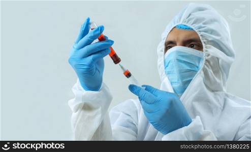 Latin doctor in bioprotective suit, face mask and blue gloves taking a blood sample in a test tube with a syringe on white background. Latin doctor in bioprotective suit, face mask and blue gloves taking a sample from a test tube with a syringe on white background