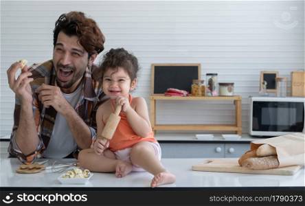 Latin family young father and little daughter play cooking in the kitchen at home. Cute girl smile and holding rolling pin in her hands while dad laughing and holding an ice cream cookie