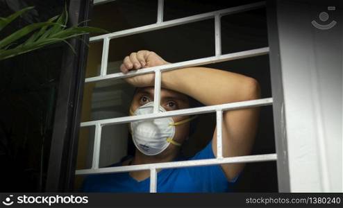 Latin man with white face mask and blue shirt inside his house looking out through the window, leaning against security bars. Latin man with white face mask inside his house looking out through the window, leaning against security bars