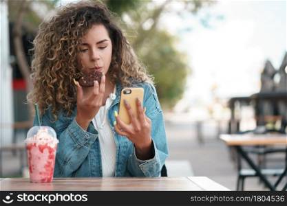 Latin woman using her mobile phone while drinking a cold drink at a coffee shop outdoors on the street. Urban concept.