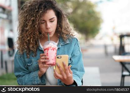 Latin woman using her mobile phone while drinking a cold drink at a coffee shop outdoors on the street. Urban concept.