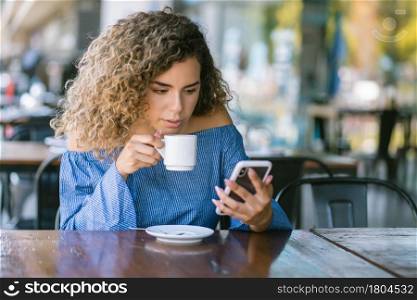 Latin woman using her mobile phone while drinking a cup of coffee at a coffee shop outdoors on the street. Urban concept.