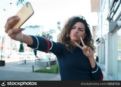 Latin young woman taking selfies with her mobile phone while standing outdoors on the street. Urban concept.