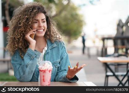 Latin young woman talking on the phone while sitting at a coffee shop outdoors on the street. Urban concept.