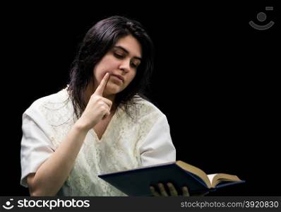 Latina teen in white dress holding a blue book with gold edging, holding a finger to her cheek and standing in front of a black background