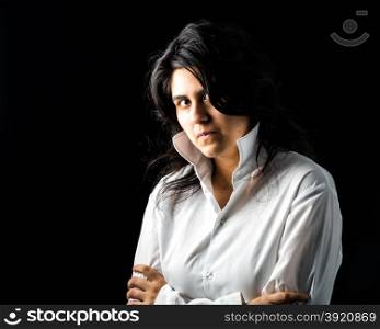 Latina teen in white long sleeved shirt standing in front of black backdrop with arms crossed and looking at the camera. Latina teen in white on black backdrop