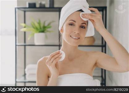 Latina woman moisturizing, cleansing facial skin, using cotton pads and micellar water or milk, removing makeup after the shower in bathroom. Self care, personal hygiene, skincare daily routine.. Woman moisturizing, cleansing facial skin, using cotton pads in bathroom. Personal hygiene, skincare