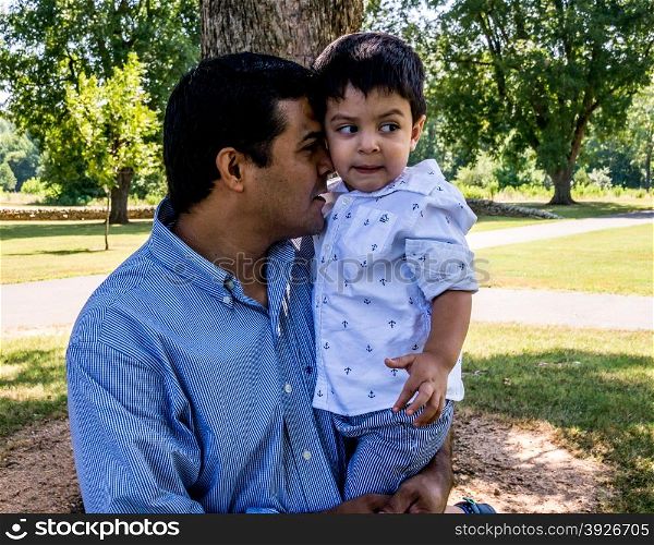 Latino father standing outside in a park with trees behind him holding his son in his arms