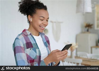 Laughing african american teen girl wear wireless earphone holds smartphone listen audio message from friend. Modern biracial female enjoy quality sound, use music apps, having fun at home.. African american teen girl in earphone hold smartphone, listen audio message, use music apps at home