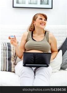 Laughing beautiful pregnant woman sitting on sofa at home with laptop and credit card&#xA;