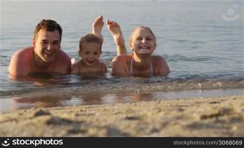 Laughing little boy flanked by his loving parents paddling together in the shallow water at the edge of the sea