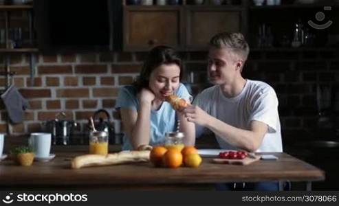 Laughing man feeding his cute brunette girlfriend croissant while having breakfast together in domestic kitchen. Loving couple enjoying nice morning and sharing meal together.