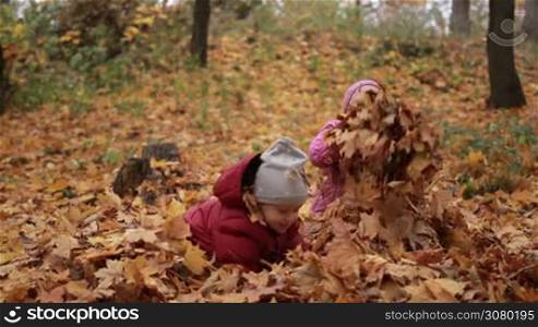 Laughing siblings throwing yellow maple leaves up in autumn park. Cute toddler boy and teenage sister sitting in pile of fall foliage and playing with autumn leaves outdoors. Joyful kids resting in autumn park. Slow motion.