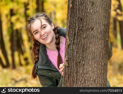 Laughing teen girl. A girl looks out from behind a tree in the park. Smiling happy face.