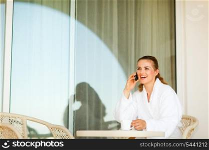 Laughing woman in bathrobe sitting at table on terrace having coffee and speaking phone&#xA;