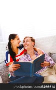 Laughing young couple sitting on couch and looking in photo album &#xA;