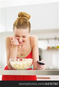 Laughing young woman eating popcorn and watching tv in kitchen
