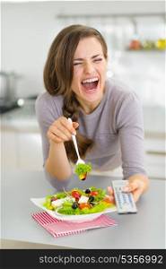 Laughing young woman eating salad and watching tv in kitchen