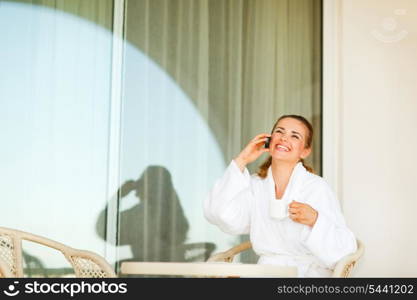 Laughing young woman in bathrobe with cup of coffee speaking mobile phone on terrace