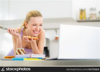 Laughing young woman spread toast with chocolate cream while studying in kitchen